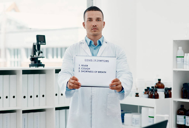 Seek help if you see any of these symptoms. Portrait of a young scientist holding a sign with a list of COVID-19 symptoms on it in a modern laboratory - Foto, afbeelding