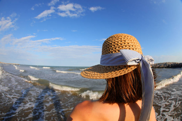 молода біла дівчина з солом'яним капелюхом дивиться на море з берега
 - Фото, зображення
