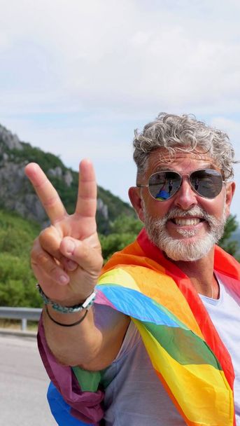 Портрет седовласого пожилого кавказца бисексуальности с бородой и солнцезащитными очками с радужным ЛГБТ-флагом и жестом мира в горах. Празднует месяц гордости, день радужного флага - Фото, изображение