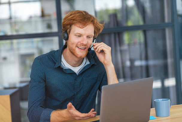 Caucasian young man customer support call center operator or receptionist sitting at the workplace in a modern office consulting a client, uses a headset, smiles friendly - 写真・画像