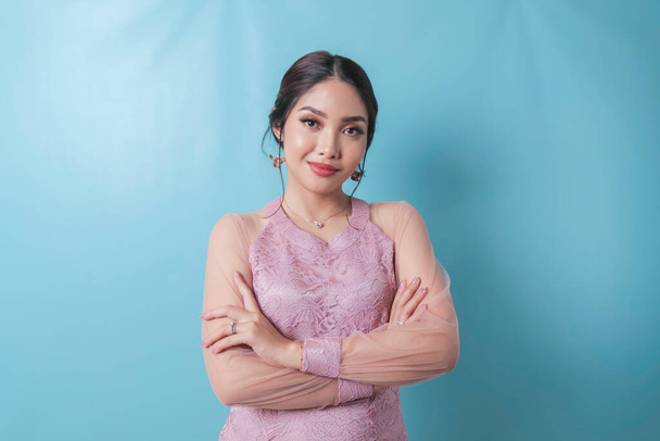 Portrait of a confident smiling girl standing with arms folded and looking at camera isolated over blue background, wearing modern kebaya dress - Fotografie, Obrázek