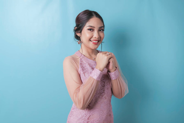 Smiling young Asian woman wearing modern kebaya gesturing Chinese greeting or Gong Xi isolated over blue background - Fotó, kép
