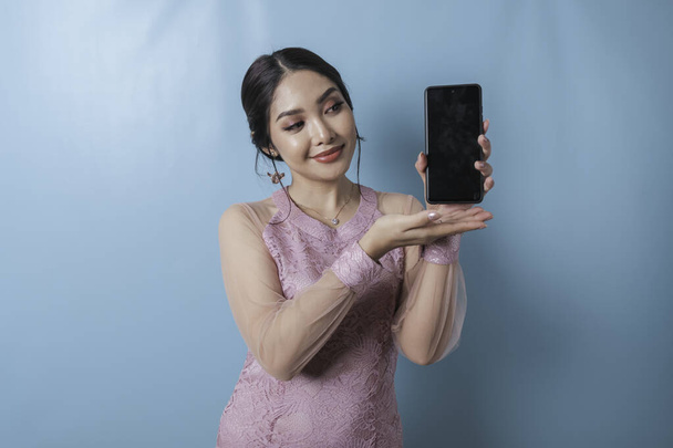 A portrait of a cheerful Asian woman showing copy space on her smartphone over blue background, wearing a modern kebaya - Fotografie, Obrázek