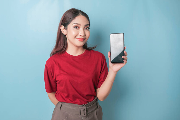 Excited Asian woman wearing a red t-shirt showing and pointing copy space on her smartphone, isolated by blue background - Φωτογραφία, εικόνα