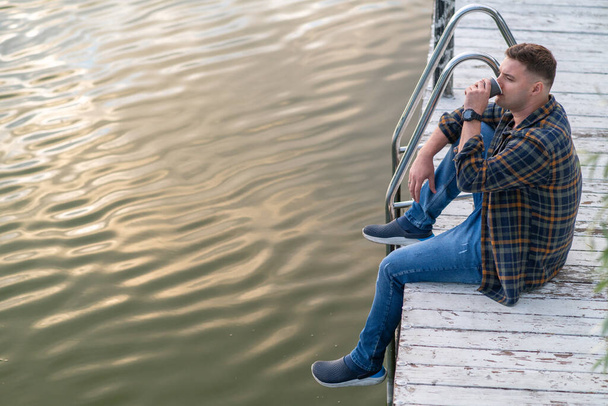 High angle view of a casual guy relaxing in a wooden deck above a tranquil lake at sunset drinking a cup of coffee - 写真・画像