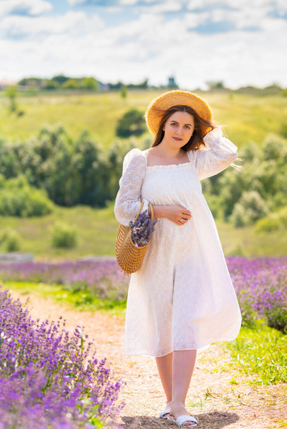 Stylish young lady walking down a rural footpath in a fresh white summer frock and straw hat between lavender fields - Фото, изображение