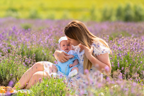Loving young mother kissing her small baby son as she relaxes sitting amongst the lavender in a farm field - Фото, зображення
