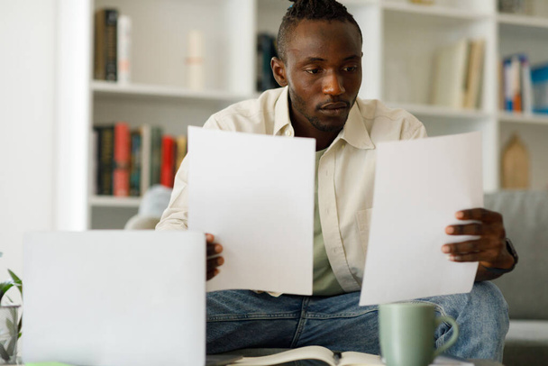Νεαρός Αφρικανός κάθεται στο τραπέζι με σύγχρονο φορητό υπολογιστή στο γραφείο του σπιτιού, διαβάζοντας γράμματα, κρατώντας έγγραφα ή λογαριασμούς, στοχαστικός τύπος πήρε άσχημα νέα, ειδοποίηση χρέους. Υψηλής ποιότητας φωτογραφία - Φωτογραφία, εικόνα