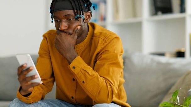 Λυπημένος νεαρός μαύρος Αμερικανός κάθεται στον καναπέ, κοιτάζοντας το τηλέφωνο,. Υψηλής ποιότητας φωτογραφία - Φωτογραφία, εικόνα
