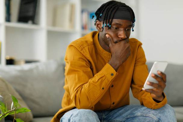 Расстроенный чернокожий африканский мужчина, читающий плохие новости в сообщении, глядя на экран телефона, сидящего на диване. Высокое качество фото - Фото, изображение