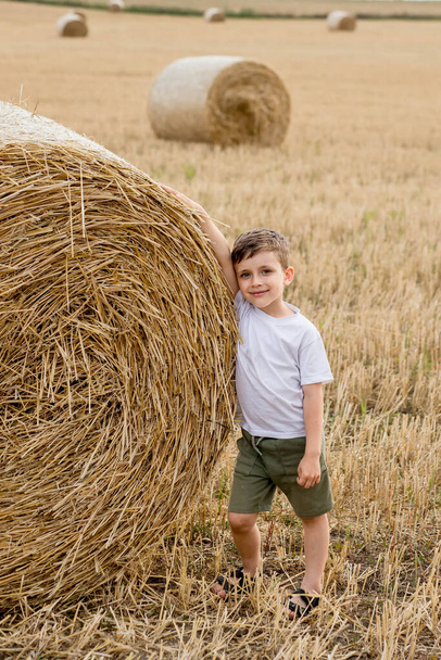 A cute little boy in a white t-shirt and khaki shorts is standing near round bales of hay. Photo session in the field. - Foto, imagen