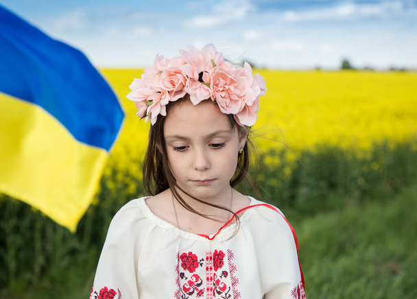 Close-up portrait of sad Ukrainian girl 7 years old in traditional embroidered blouse with blue-yellow flag on background of yellow blooming rapeseed field. Children against war. Support Ukraine - 写真・画像
