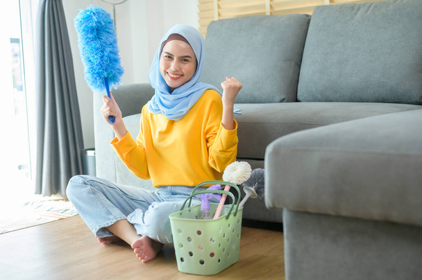 A Young happy muslim woman wearing yellow gloves  and holding a basket of cleaning supplies in living room. - Φωτογραφία, εικόνα