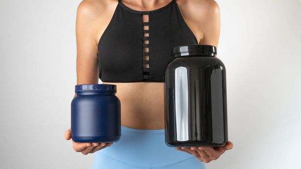 Athletic slender woman holding cans of protein and sports supplements for an effective workout in the gym. High quality photo - Foto, afbeelding