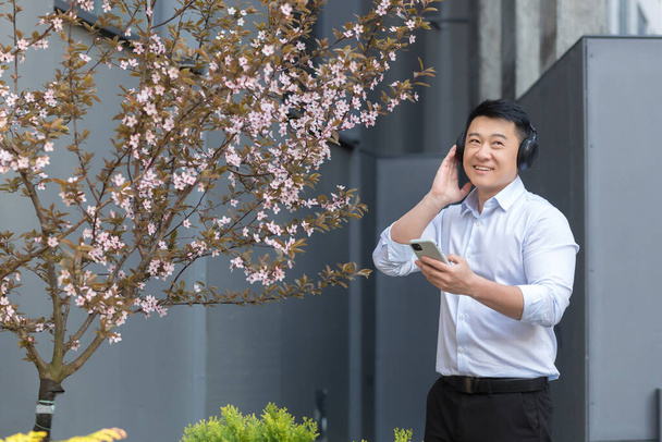 Cheerful and smiling successful Asian business man listening to music using smartphone and big headphones, manager resting outside office in shirt - Φωτογραφία, εικόνα