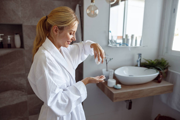 Young beautiful Caucasian woman pouring body lotion on hand in bathroom. Anti-aging concept. At home - Zdjęcie, obraz