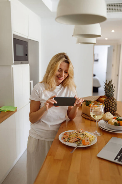 Блондинка фотографирует тарелку с креветками, макаронами и белым вином. Блогер еды. Вертикаль. - Фото, изображение