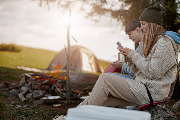 Caucasian females sitting at fire in camp site and using phones when resting. Travelers. Resting. - 写真・画像
