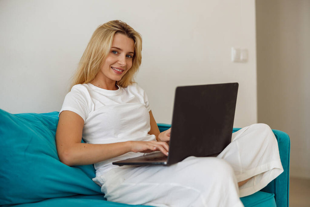 Portrait of blonde pretty young woman smiling to camera while sitting on sofa with laptop. Homey job - 写真・画像