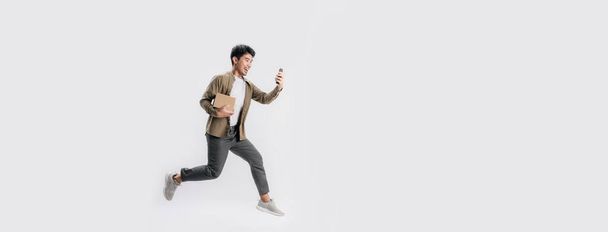 Πλήρες μήκος όμορφος Ασιάτης άνδρας κρατώντας κουτί και να χρησιμοποιήσετε το smartphone που τρέχει και πηδώντας στον αέρα σε πανό λευκό φόντο. Νεαρό αρσενικό χαρούμενος και τη χρήση του κινητού τηλεφώνου με ψώνια σε απευθείας σύνδεση έννοια. - Φωτογραφία, εικόνα
