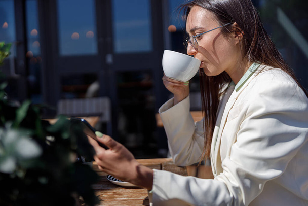 Atrakcyjna kobieta biznesu w okularach przeciwsłonecznych pije kawę siedząc w kawiarni i odwracając wzrok - Zdjęcie, obraz
