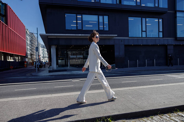 Beautiful woman in trendy outfits crosses road against backdrop of city buildings and looking away - Fotoğraf, Görsel