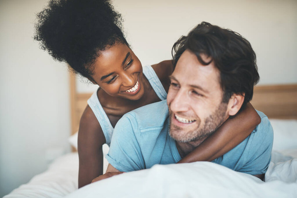 Interracial, playful and loving couple cuddling in bed and enjoying the weekend together at home. Affectionate, happy and bonding diverse lovers embracing and having fun in their bedroom. - Фото, зображення
