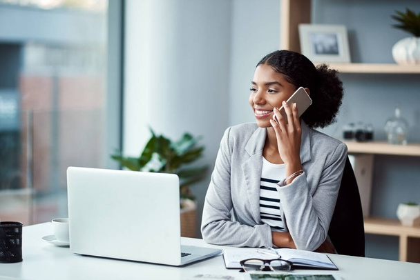 Business woman talking on phone call or young entrepreneur answering cellphone in front of laptop in work office. Happy, African American female smiling and receiving good news while sitting at desk - 写真・画像