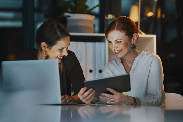 Now we can stay connected throughout the night. two businesswomen using a digital tablet together in an office at night - Photo, Image