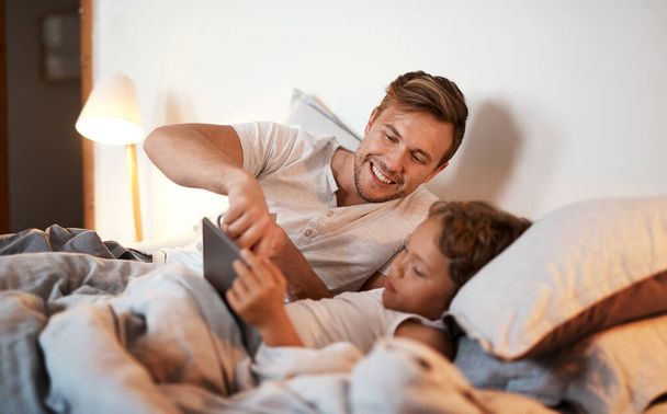 Ξέρω την απάντηση σ 'αυτό. ένα νεαρό αγόρι που χρησιμοποιεί ένα ψηφιακό tablet ενώ βρίσκεται στο κρεβάτι με τον πατέρα του - Φωτογραφία, εικόνα