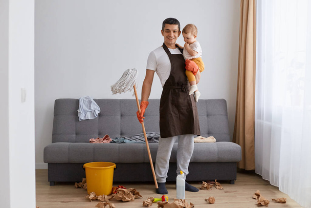 Portrait of satisfied smiling man wearing casual clothing and brown apron cleaning house with baby daughter in hands, holding mop, looking at camera with positive expression, doing housework. - Photo, Image