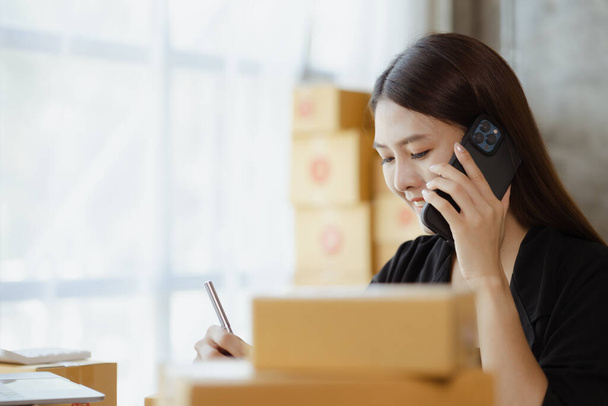 Asian woman is talking on the phone with a customer to confirm an order, she owns an online store, she packs and ships through a private transport company. Online selling and online shopping concepts. - Fotografie, Obrázek