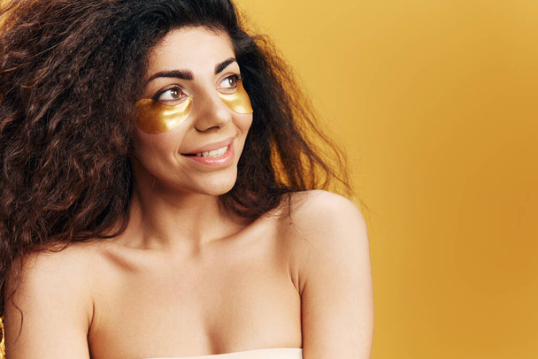 Hymyilevä onnellinen melko ruskettunut latinalaisamerikkalainen nainen, jolla on kultaiset hydrogeelilaastarit silmän alla, katso sivuun aiheuttaen eristettyä keltaisella taustalla. Kosmeettinen tuote mainos Natural kauneus käsite Studio muotokuva Kopioi tilaa - Valokuva, kuva