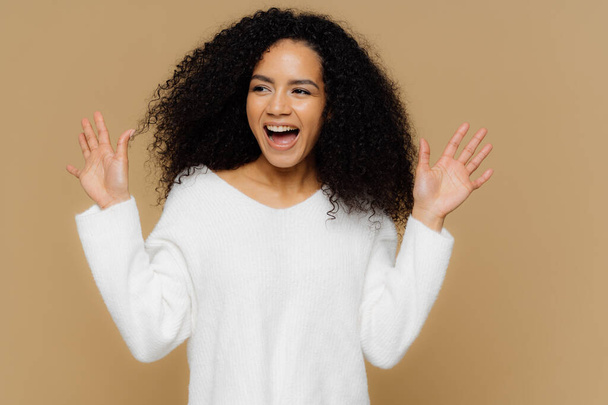 Glad overemotive woman surprised by pleasant relevation, raises hands and shows palms, focused aside, wears casual white sweater, models over brown background. Positive human emotions and feelings - Фото, зображення