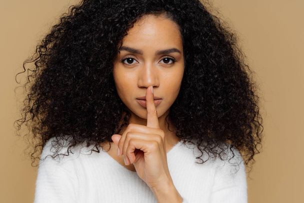 Image of serious confident dark skinned woman makes shush gesture, keeps fore finger over lips, tells secret information, asks not spread rumors, dressed in casual outfit, isolated on beige background - Foto, Imagen