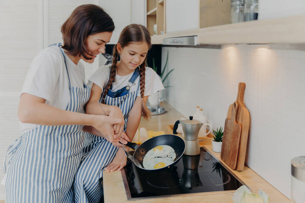 Busy mother and daughter wear striped aprons, pose at kitchen near cooker, fry eggs on pan, prepare fast breakfast, enjoy domestic atmosphere. Mum teaches small kid to cook. Happy family concept - Photo, image