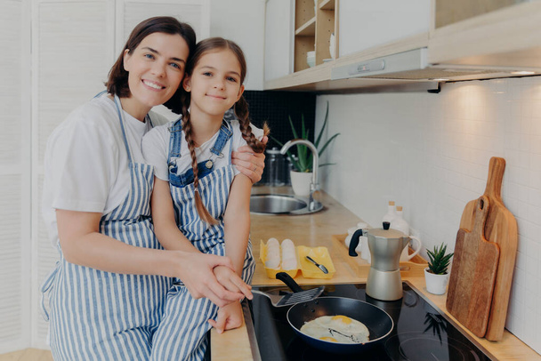 Lovely mother and daughter in aprons embrace and smile happily, fry eggs on modern stove in kitchen, use frying pan, prepare tasty breakfast. Family, children, motherhood and cooking concept - Foto, Imagen