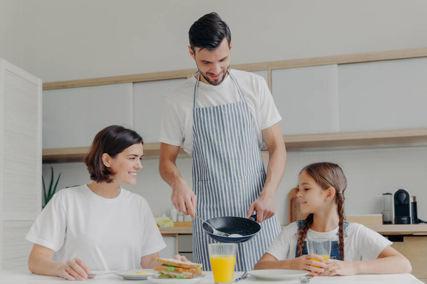 Busy caring father prepares delicious breakfast for wife and daughter, wears apron, puts fried eggs on plate. Cheerful mother and child talk to each other while sit at kitchen table, have meal - 写真・画像