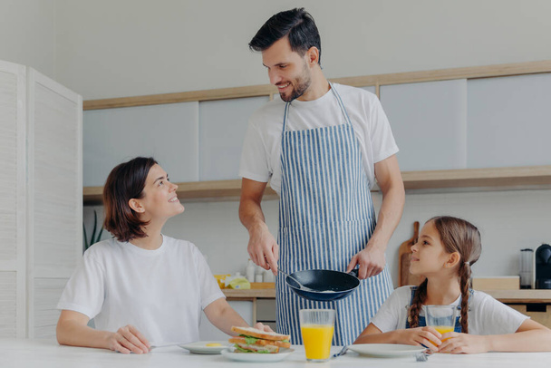 Happy father, mother and daughter gather together at kitchen, eat delicious breakfast, dad prepared fried eggs, being in good mood, ready to start new day. Lovely family enjoy tasty meal at home - Foto, immagini