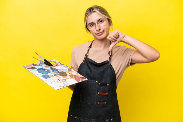 Young artist caucasian woman holding a palette isolated  on yellow background showing thumb down with negative expression - Foto, Bild