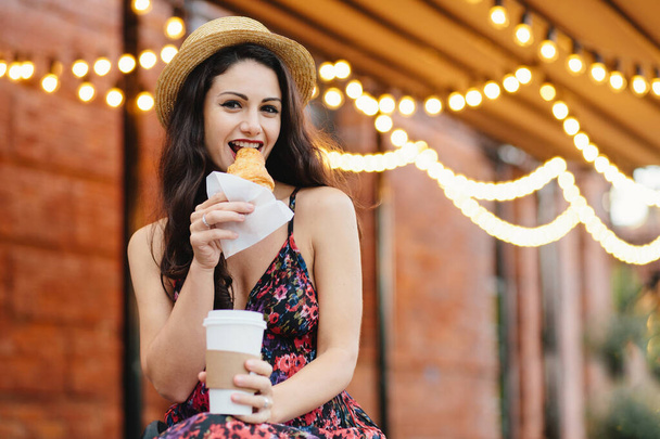 Pretty woman with dark hair, shining eyes eatting with pleasure tasty croissant and drinking takeaway coffee while sitting at cozy balcony. Young woman in summer hat and dress relaxing at cafe - Photo, Image