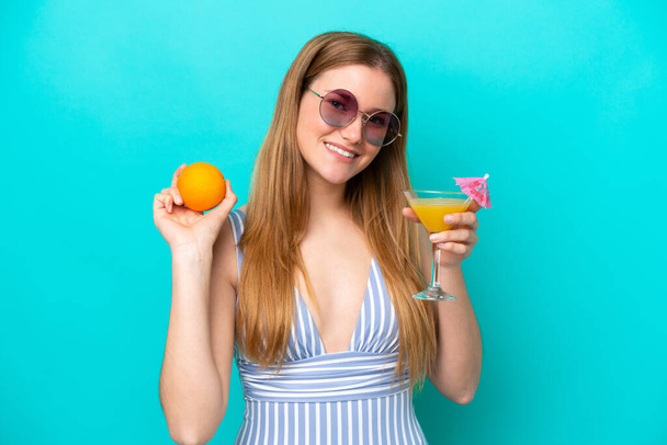 Young woman isolated on blue background in swimsuit and holding a cocktail - Photo, Image