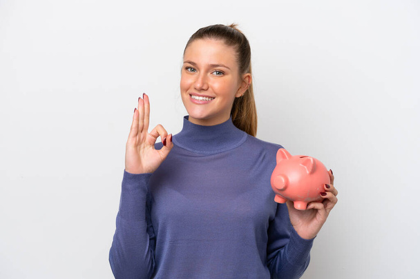 Young caucasian woman holding a piggybank isolated on white background showing ok sign with fingers - Fotografie, Obrázek