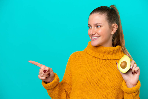 Young caucasian woman holding an avocado isolated on blue background pointing to the side to present a product - Foto, immagini