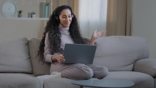 Joyful successful hispanic girl sit on sofa make video call communicate remote in online conference use laptop and headphones happy smiling young lady freelancer working at home concept modern devices - Materiał filmowy, wideo