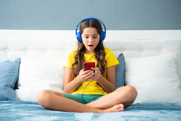 Teenager child girl wearing headphones watching videos on smart phone sitting on bed in her room. Surprised face, surprise emotions of teenager girl - Fotografie, Obrázek