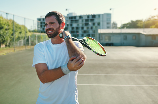 Tennis elbow can be caused by repetitive wrist and arm motions. a sporty young man holding his elbow in pain while playing tennis on a tennis court - Φωτογραφία, εικόνα