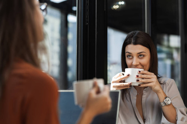 Zwei junge Geschäftsfrauen in einem Café, die sich eins zu eins treffen. Freunde nach der Arbeit quatschen und trinken an einem sonnigen Tag Kaffee am Fenstertisch. - Foto, Bild