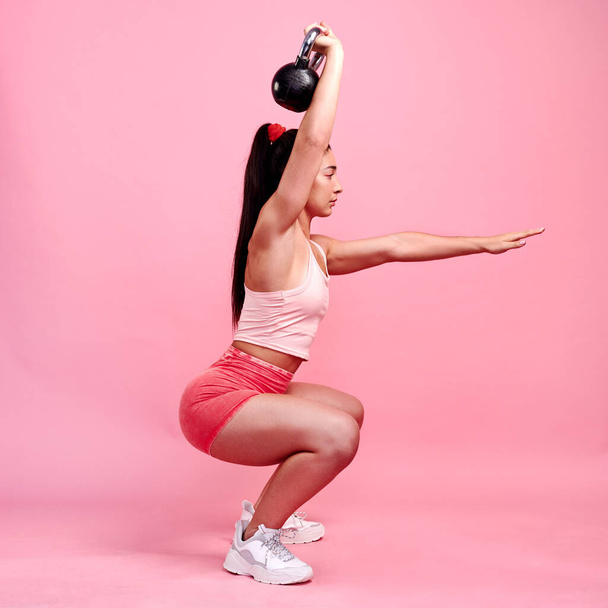 Fully focused on her form. Studio shot of a sporty young woman doing squats with a kettlebell against a pink background - 写真・画像