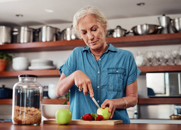 These days, she only eats healthy. an attractive senior woman chopping up apples and other fruit while preparing breakfast in the kitchen - Foto, imagen